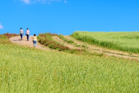 SIENA, ITALY - MAY 21, 2017 - Three people walk along a dirt road surrounded by green fields and...