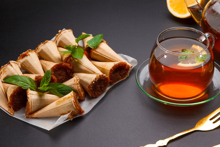 Homemade honey wafers rolled into a cone, filled with condensed milk, a cup of tea and lemon on a black tableの写真素材