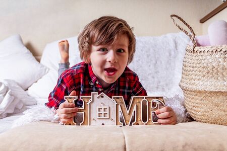 Surprised funny little curly kid boy with smiley face lying on the bed at home. Stay at home concept due quarantine coronavirus COVID-19 pandemic prevention. Wooden house inscription home.の写真素材