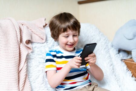 Happy little child boy playing online game, watching video on cellphone, sitting on sofa...