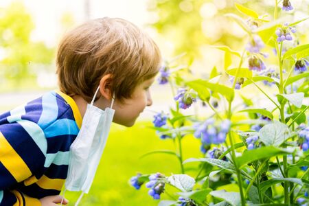 Happy kid boy taking off his medical protective mask and breathing air, smelling flowers. Cute child stay outdoors in park at sunny day. End of quarantine, covid-19 exit pandemic conceptの写真素材