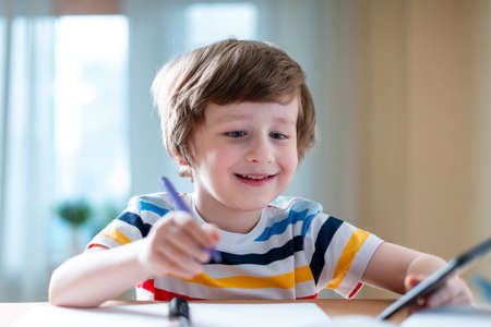 Back to school. Distance learning online education. Caucasian smile kid boy studying at home with smartphone and doing school homework. Smiling child siting at table with cell phone.の写真素材
