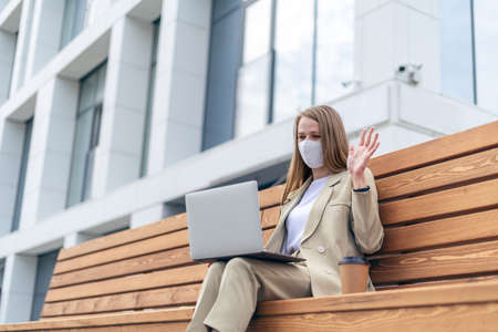 Beautiful business woman in medical protective mask and having a video call at laptop sit down on bench outside on a urban city street. Happy lady girl distance learning and online education.の写真素材