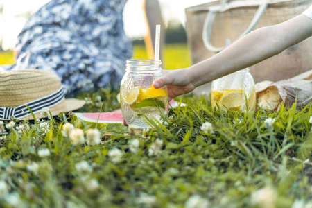 Child holding yellow and green glass with cold lemonade on summer picnic on the grass with family. The boy reaches for a glass of juice with lemon and lime. Selective focus, blurred backgroundの写真素材