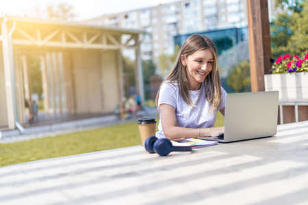 Woman with headphones having a video call with laptop outside in park. Happy and smiling girl working and drinking coffee. Using computer. Distance learning online education and online shops.の写真素材