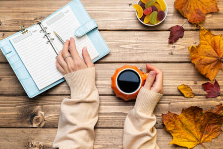 Top view composition with woman's hands in sweater, pumpkin styled cup of coffee and autumn themed decoration, fallen leaves and notebook on wooden background. Flat lay, copy space.の写真素材