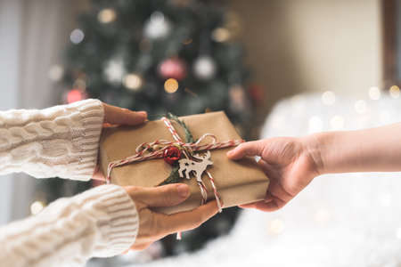 Woman in sweater giving a wrapped Christmas gift box to child. Glowing snow bokeh, fir tree. Winter...