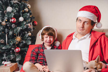 Family in Santa hat, father and crying child boy with laptop sitting on a couch in the living room with dog.の写真素材