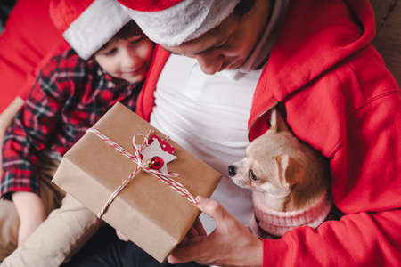 Happy family in Santa hat, father and child son giving Christmas gift at home. Sitting on a couch in the living room with puppy dog in sweaterの写真素材