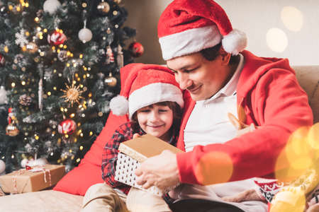 Family in Santa hat, father and child son open Christmas gift at home. Sitting on a couch in the living room with puppy dog in sweaterの写真素材