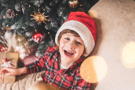 Cute little laughing boy in red hat waiting Santa Claus in living room with Christmas tree at homeの写真素材