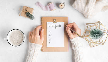 Desktop of woman hands with blank notepad writing 2021 goals text. Flat lay of white working table...