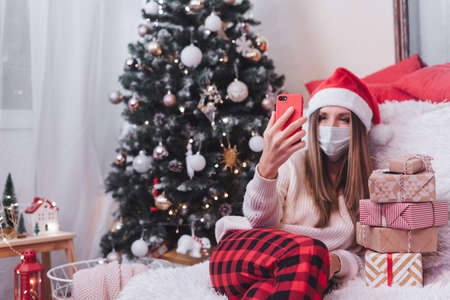 Woman in medical protective mask with gift box having a video call chat at smartphone, enjoy Christmas holidays at home with Christmas treeの写真素材