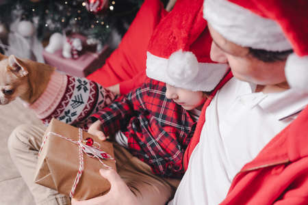 Family in Santa hat, father and child son open Christmas gift at home. Sitting on a couch in the living room with puppy dog in sweaterの写真素材