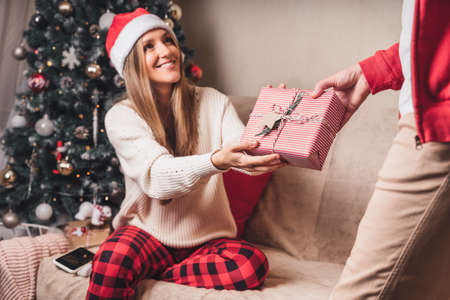 Surprising her... Close-up rear view of man in red sweater holding a Christmas gift box behind his back while standing near decorated christmas treeの写真素材