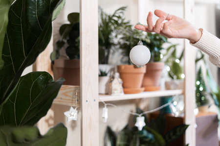 Women hands with Christmas toy ball among plants, decoration and lightsの写真素材