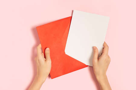 Red paper envelope with blank white note mockup in woman hands, inside on pink pastel background. Flat lay, top view. Romantic love letter for Valentines day concept.の写真素材