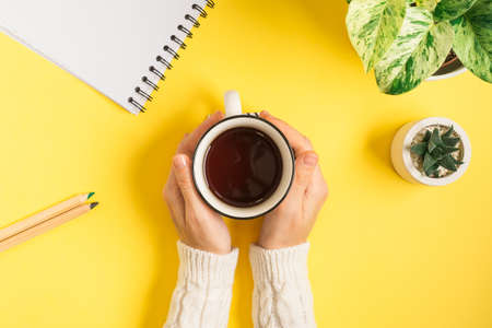 Desktop of woman hands with cup of tea. Flat lay of yellow working table background with notepad, stationery and plants . Top view, mock up greeting card, craft Notebook and pen.の写真素材