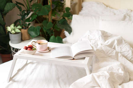 Breakfast in bed. Heart shaped white plate with fresh strawberries, cup of coffee, book and plants...
