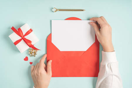 Flat lay of woman hands holding red paper envelope with blank mockup greeting card. Blue working table background with Valentine gift, letter, heart shape and decoration. Top view, mock up.の写真素材