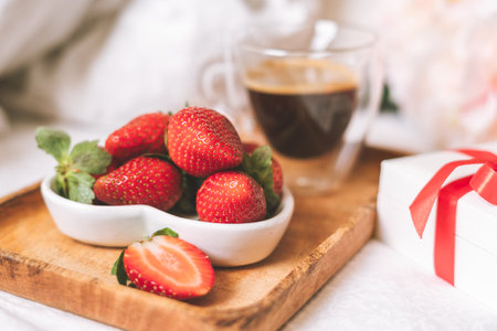 Breakfast for Valentines Day. Heart shaped white plate with fresh strawberries, cup of coffee, gift and flowers with gift in bed. Still life composition.の写真素材