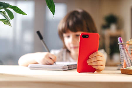 Back to school. Distance learning online education. Caucasian smile kid boy studying at home with mobile smart phone and doing school homework. Thinking child siting at table with cell phoneの写真素材