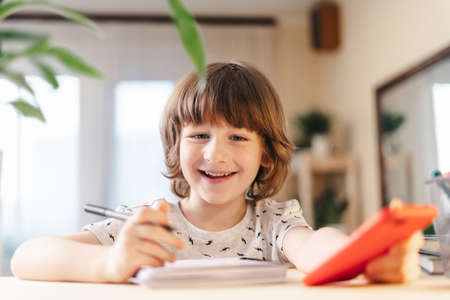 Back to school. Distance learning online education. Caucasian smile kid boy studying at home with mobile smart phone and doing school homework. Thinking child siting at table with cell phoneの写真素材