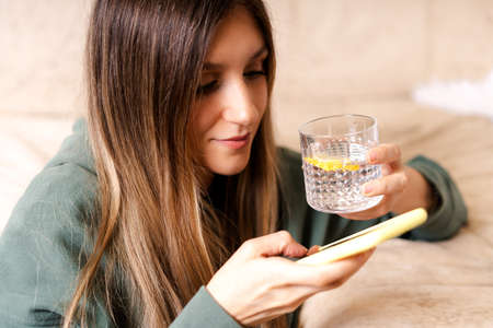 Distance learning online education and work. Beautiful woman in hoody texting and chatting at mobile cell phone, relax from home, sitting on the couch holding glass of water.の写真素材