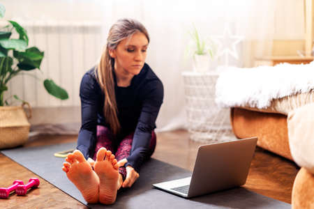 Young sporty attractive woman working out practicing yoga and using laptop and dumbbells at home in...