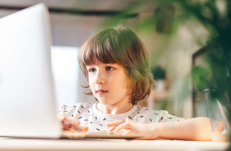 Distance learning online education. Caucasian smile kid boy studying at home with laptop and doing school homework. Thinking child siting at table with notebook. Back to school.の写真素材