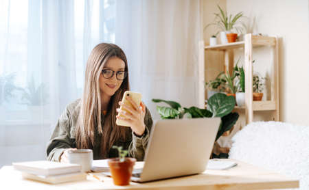 Freelance woman in glasses with mobile phone typing at laptop and working from home office with...