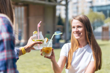 Two beautiful young women sitting on the grass and laughing while having a lemonade drink in glass jars, during a friendly hang out in city park.の写真素材