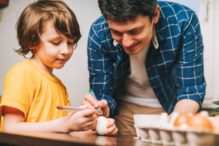Easter day. Father and son painting eggs with crayons on wooden background. Family sitting in a kitchen. Preparing for Easter, creative homemade decoration.の写真素材
