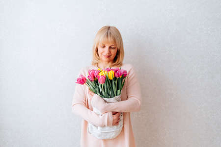 Portrait of Beautiful mature happy smiling woman with blonde hair, holding bouquet of pink and yellow tulip flowers in knitted bag. White background, copy space, close up.の写真素材