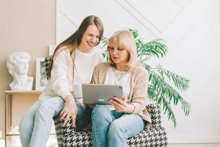 Mature senior woman with laughing adult daughter using digital tablet computer device. Caucasian...