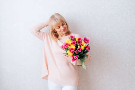 Portrait of Beautiful mature happy smiling woman with blonde hair, holding bouquet of pink and yellow tulip flowers in knitted bag. White background, copy space, close up.の写真素材