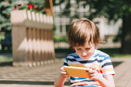 A beautiful child sits on the wooden playground and looks at the mobile cell phone in the summer....