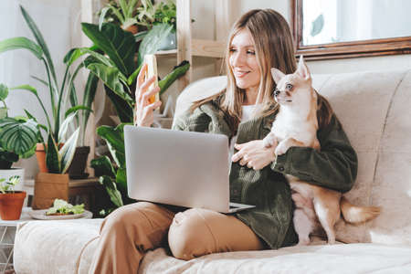 Freelance woman taking selfie with mobile phone and dog, typing at laptop and working from home office. Happy girl sitting on couch in living room with plants. Distance learning and online education.の写真素材