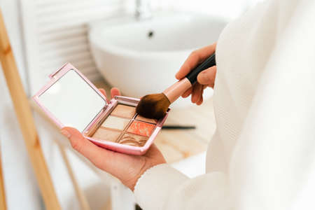 Close up of cropped caucasian woman with long hair holding powder palette and brush, doing makeup...
