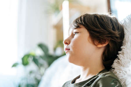 Portrait of thinking sad child Kid boy in green t shirt playing video games and watching tv in living room at sunny day. Stylish interior with a lot of plants at home. Spring time.の写真素材