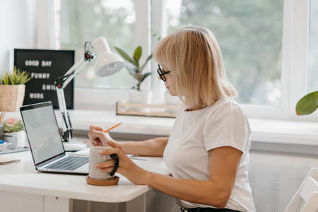 Business adult mature woman typing on laptop computer while sitting at home workplace and writing in...