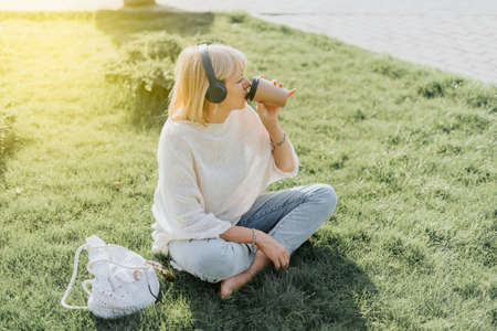 Adult Woman in headphones sitting on the grass outside in summer park at sunset. Happy and smiling senior listening music and drinking coffee in paper cup. Relax and meditation.の写真素材