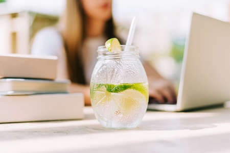 Beautiful young student girl in the summer sunny city park drinking a cocktail with lemon from glass jars. Woman sits and typing at the laptop. Selective focus.の写真素材