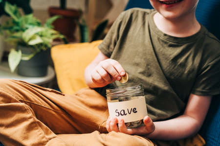 Close up of Little child kid boy hands grabbing and putting stack coins in to glass jar with save...