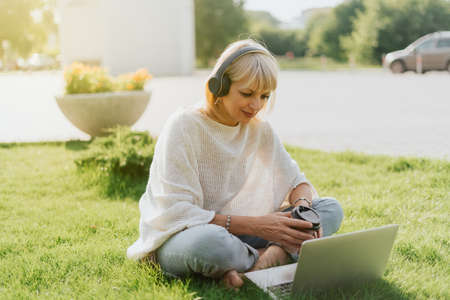 Mature adult Woman in headphones using laptop, sitting on the grass outside in park. Happy and...