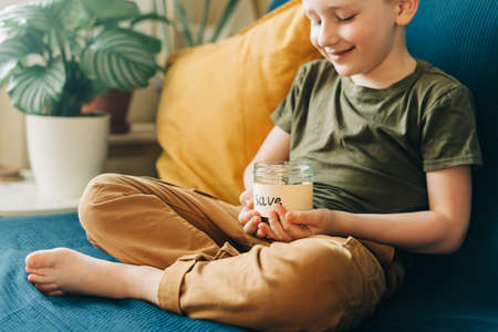Close up of Little child kid boy hands grabbing and putting stack coins in to glass jar with save label. Donation, saving money, charity, family finance plan concept.の写真素材