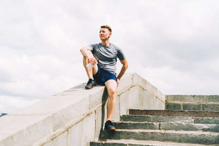 Tired Young man runner sitting on stairs and relaxing after sport training. Holding water bottle...
