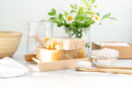 Natural bathroom and home spa tools. Zero waste sustainable lifestyle concept. Natural soap bar in wooden soap dish, cotton pads and swabs, flowers, bamboo toothbrush. White background, copy spaceの写真素材