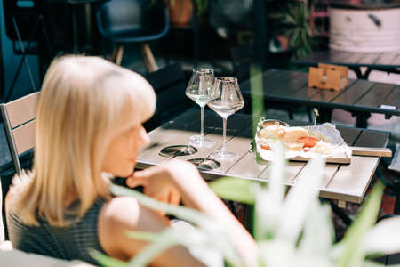 Young woman sitting in cafe outdoors with glasses and blurry restaurant background scene, drinking white wine and eating cheese. Summer sunny day on patio.の写真素材
