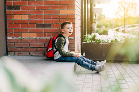 Back to school. Cute child with backpack, holding notepad and training books. School boy pupil with bag. Elementary school student going to classes. Kid sitting on stairs outdoors at brick wall.の写真素材